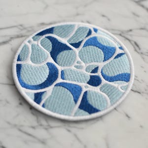 Peut inclure: Écusson rond brodé avec un motif abstrait bleu et bleu clair, bordé de blanc. Le patch a une bordure blanche et est destiné à la décoration, comme sur les vêtements ou les accessoires.