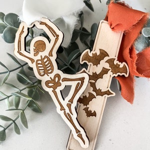 May include: Two wooden Halloween-themed bookmarks. One features a skeleton in a dancing pose, the other has bats. Both are on a white surface with fabric.