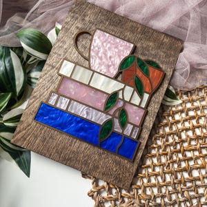 May include: Wooden plaque featuring a layered design of books, a teacup, and a potted plant. The books have blue, pink, and white sections. The teacup is pink, and the plant pot is orange. Green leaves are interspersed throughout the design.