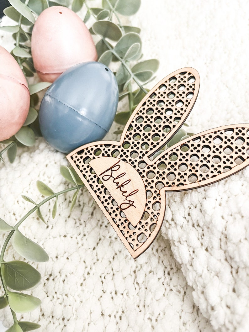 Op de afbeelding: Houten konijnenoor-kaartjeshouder met de naam "Blakeley" erop gegraveerd. De kaartjeshouder ligt op een wit, pluche oppervlak met twee pastelkleurige eieren.