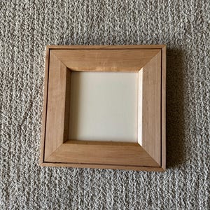 May include: A square wooden picture frame with a light brown natural wood grain finish. The frame features a wide, flat border surrounding a plain, off-white square center panel. The construction shows mitered corners and a recessed inner edge.
