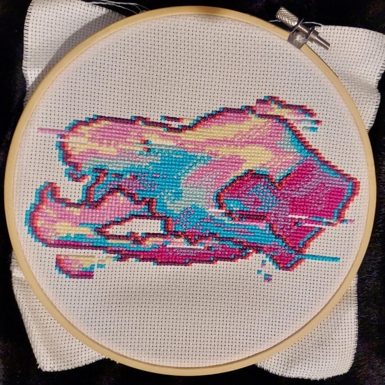 May include: A colourful abstract design embroidered on white fabric in a circular embroidery hoop. The design features shades of pink, blue, yellow, and purple.