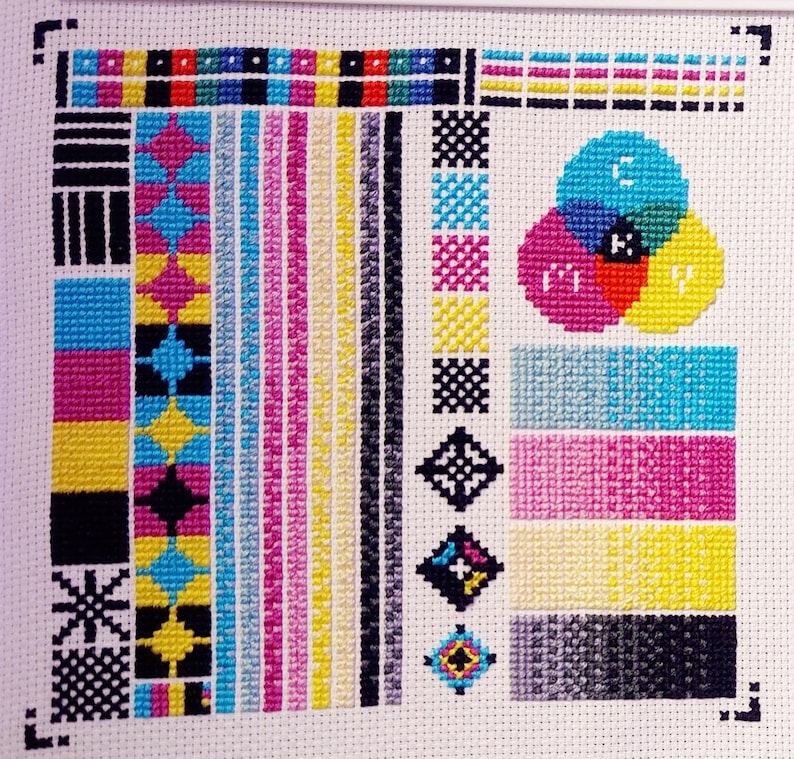 Pu&ograve; includere: Uno schema di punto croce che presenta una ruota dei colori con le lettere CMYK al centro, circondata da vari quadrati e linee colorate. Lo schema &egrave; ricamato su tessuto bianco.