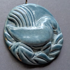 Può includere: Decorazione murale in ceramica blu con un design a cigno. Il pezzo ovale presenta una finitura lucida e un anello in ottone per appenderlo. Il cigno è raffigurato in posizione arricciata, con dettagli di piume e foglie.