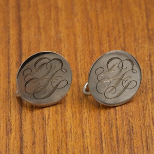 Puede incluir: Un par de gemelos de plata con un diseño de monograma. Los gemelos son redondos y tienen un acabado liso y pulido.