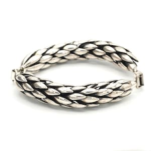 May include: A silver bracelet featuring a repeating pattern of overlapping, leaf-shaped or scale-like segments. The metal has a polished finish with darkened recessed areas between the individual links, creating a textured, dimensional appearance.