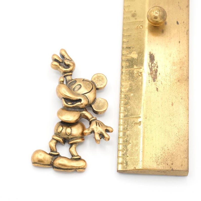 Peut inclure: Figurine dor&eacute;e de Mickey Mouse en position debout, une main lev&eacute;e en signe de paix. La figurine est &agrave; c&ocirc;t&eacute; d'une r&egrave;gle en laiton. La figurine de Mickey Mouse mesure environ 5 cm de haut.