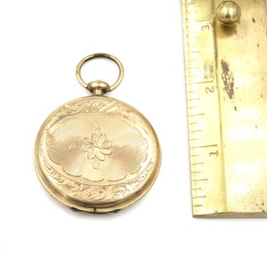 May include: A gold-colored pocket watch with an oval floral design on the front. The watch has an attached ring at the top and is next to a ruler. The watch's face has a flower in the center and is surrounded by an engraved pattern.