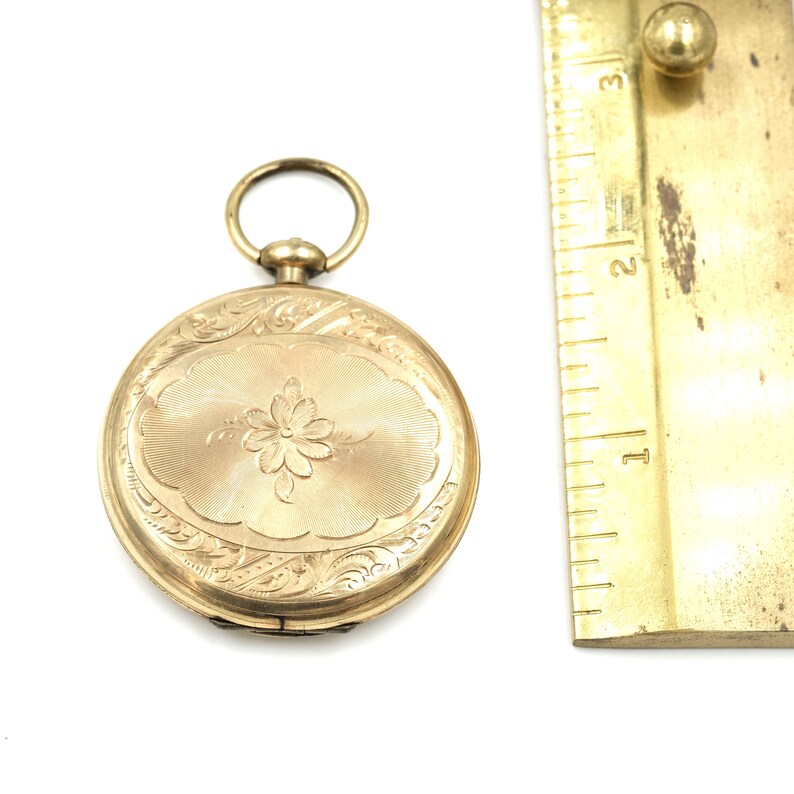 May include: A gold-colored pocket watch with an oval floral design on the front. The watch has an attached ring at the top and is next to a ruler. The watch's face has a flower in the center and is surrounded by an engraved pattern.