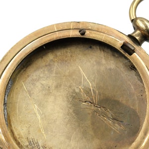 May include: Close-up of an antique pocket watch, showcasing the interior of the watch case. The watch has a brass or gold-toned metal finish, with visible scratches and wear on the surface. The watch's design includes a loop for a chain.