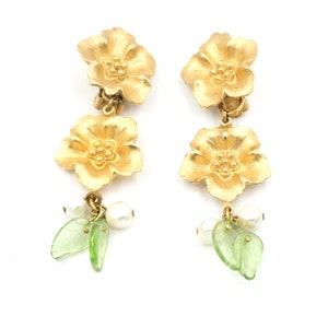 Op de afbeelding: Gouden oorbellen met bloemenhangers, groene bladeren en witte parel accenten.