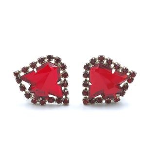 May include: A pair of red rhinestone earrings with a geometric design. The earrings are made of silver metal and have a red crystal in the center.