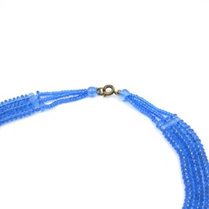Op de afbeelding: Een meerrijige ketting gemaakt van kleine, lichtblauwe glazen kralen. De ketting heeft een eenvoudige, messingkleurige sluiting.