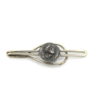 May include: Silver tie clip with a round, silver-toned coin featuring a profile of a woman with a laurel wreath. The coin is set in a silver-toned, teardrop-shaped frame.