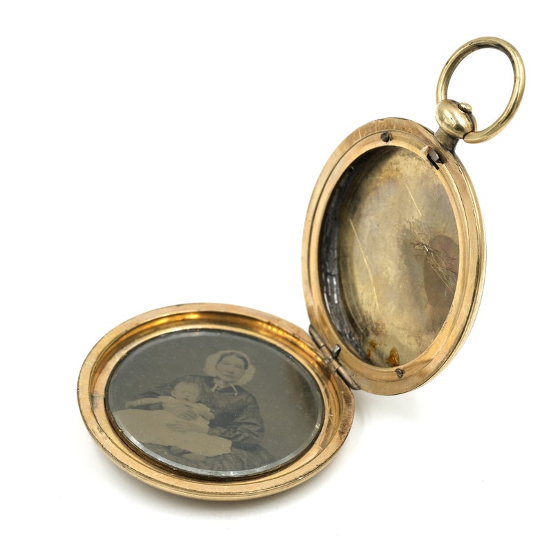 May include: Open gold-colored oval locket with a small loop at the top. The locket is open, revealing a black and white photograph of a person holding a baby. The locket is a vintage piece, likely for holding a cherished memory.