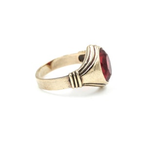 May include: A gold ring with a red gemstone set in a bezel setting. The ring has a simple band and a slightly raised setting.