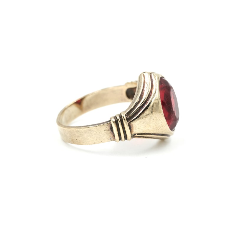 May include: A gold ring with a red gemstone set in a bezel setting. The ring has a simple band and a slightly raised setting.