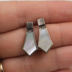 May include: A pair of silver-toned earrings with a geometric design. Each earring features a rectangular top and a pearlescent, diamond-shaped pendant. The earrings are displayed against a neutral background.