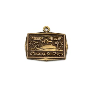 May include: A vintage gold-toned metal pendant with the text "AL BAHR W.S.A. TEMPLE" and "Oasis of San Diego." The pendant features a depiction of palm trees, a building, and a stylized bird or fish.