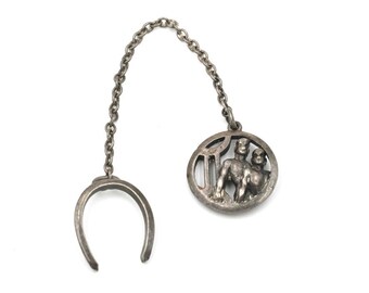 May include: A silver-toned tie clip featuring a horseshoe-shaped clasp connected by a chain to a circular pendant. The pendant depicts two figures, possibly representing a zodiac sign, within a decorative frame. The design is detailed and vintage.