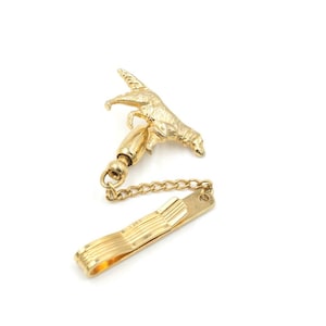 May include: A gold-toned tie clip featuring a detailed dog figurine, a chain, and a clasp. The dog is sculpted in a dynamic pose, and the clip has a ribbed design. The clip is designed to secure a tie to a shirt.