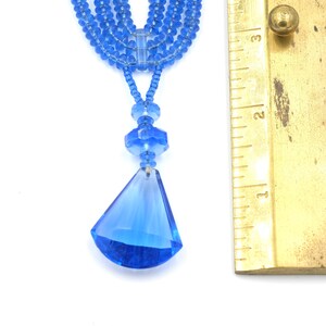Op de afbeelding: Een meerstrengs ketting met een blauwe kristallen hanger. De ketting is gemaakt van kleine, gefacetteerde glazen kralen. De hanger heeft een traanvorm met een puntige punt. De ketting is ongeveer 5 cm lang.