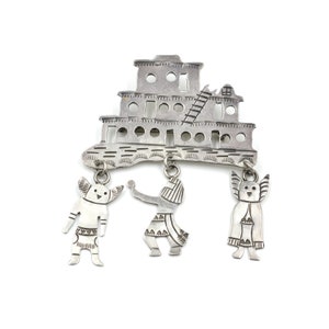 May include: A silver brooch featuring a stylized depiction of a pueblo building with three dangling figures. The figures are stylized representations of people with wings and geometric patterns.