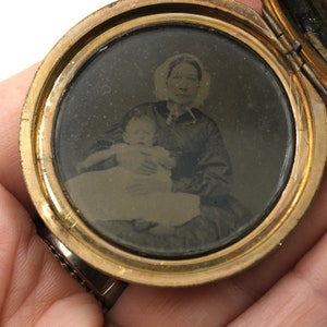 May include: A vintage gold-toned locket, open to reveal a sepia-toned photograph. The photograph depicts a woman holding a baby. The locket's circular frame surrounds the image, with visible wear and tear.