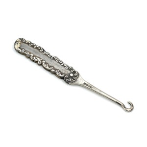 May include: A silver hook tool with an ornate handle. The handle is decorated with a swirling pattern and the tool is marked "STERLING".