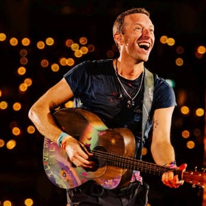 May include: Acoustic guitar with a colorful painted design, held by a person wearing a dark blue t-shirt. The guitar has a rainbow-colored design on the body and the words "I LOVE WH" on the wristband. The background is blurred with golden lights.