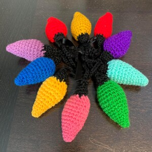 Puede incluir: Un conjunto de nueve adornos de bombilla de luz de ganchillo en varios colores, incluyendo morado, rosa, azul, verde, amarillo y rojo. Los adornos están conectados por hilo negro.