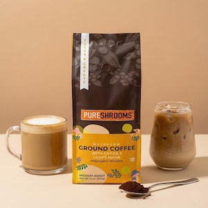 May include: A bag of PURESHROOMS mushroom ground coffee with Chaga and Lion's Mane, medium roast, net weight 340g. A clear glass mug with coffee and foam, a glass of iced coffee, and a spoon with coffee grounds are also in the image.