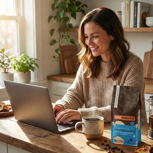 May include: A woman sits at a wooden table with a laptop, a mug of coffee, and a bag of PURESHROOMS ground coffee. The coffee bag is blue and brown with the text "Mushroom Ground Coffee with Ashwagandha & Chaga". Coffee beans are scattered on the table.