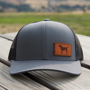 May include: A gray and black trucker hat with a brown leather patch featuring a silhouette of a dog. The hat has a mesh back and a curved brim.