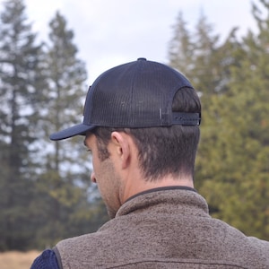 Puede incluir: Gorra de camionero azul marino con parte trasera de malla y visera plana. La gorra tiene un cierre a presi&oacute;n para un ajuste ajustable. La persona de la imagen lleva un chaleco marr&oacute;n y una camisa azul.