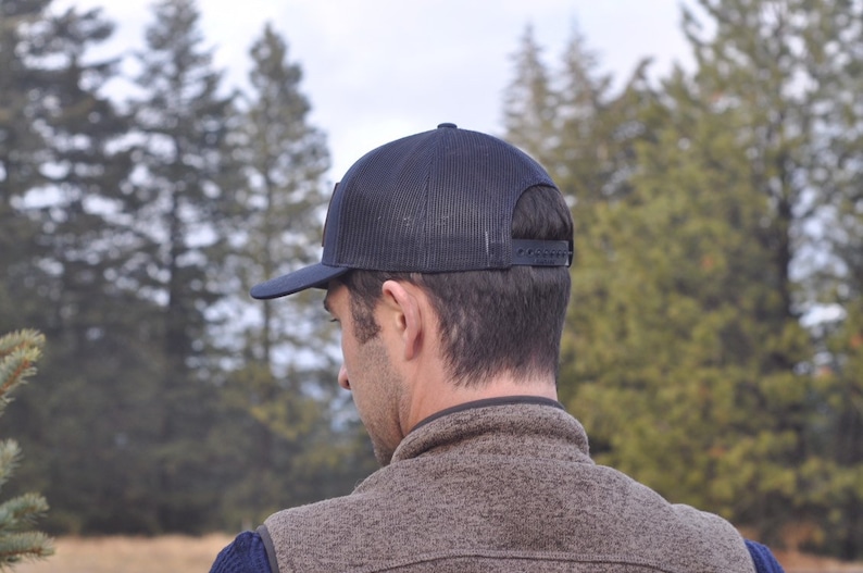 Puede incluir: Gorra de camionero azul marino con parte trasera de malla y visera plana. La gorra tiene un cierre a presi&oacute;n para un ajuste ajustable. La persona de la imagen lleva un chaleco marr&oacute;n y una camisa azul.
