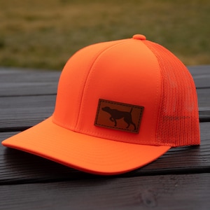 May include: Bright orange trucker hat with a mesh back and a curved brim. A brown leather patch on the front displays a silhouette of a dog. The hat is designed for outdoor activities and offers sun protection.