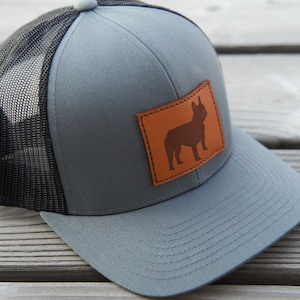 Puede incluir: Gorra trucker gris con parte trasera de malla negra y visera curva. Un parche de cuero marr&oacute;n en la parte delantera muestra la silueta de un bulldog franc&eacute;s. La gorra tiene cierre snapback.