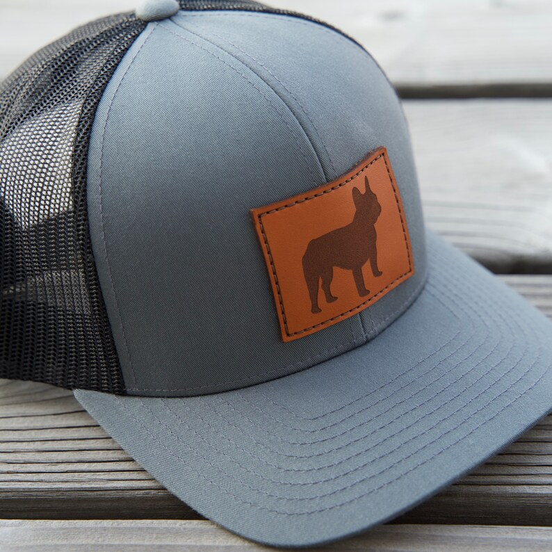 Puede incluir: Gorra trucker gris con parte trasera de malla negra y visera curva. Un parche de cuero marr&oacute;n en la parte delantera muestra la silueta de un bulldog franc&eacute;s. La gorra tiene cierre snapback.