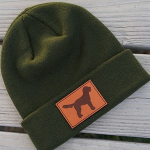 May include: A dark green beanie with a brown leather patch featuring a silhouette of a dog. The patch is stitched around the edges.
