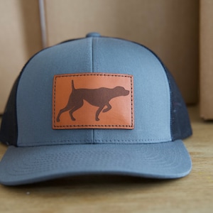 May include: Gray trucker hat with a brown leather patch featuring a silhouette of a dog. The hat has a gray front panel, a black mesh back, and a curved brim. The dog silhouette is in a running pose.