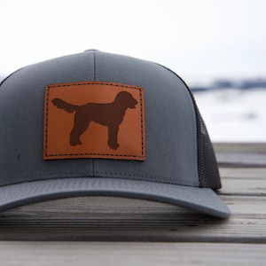 May include: A gray and black trucker hat with a brown leather patch featuring a silhouette of a dog.