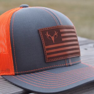 K&ouml;nnte beinhalten: Eine graue und orangefarbene Trucker-Kappe mit einem braunen Leder-Patch, der eine amerikanische Flagge und eine Hirsch-Silhouette zeigt. Die Kappe hat ein Netzr&uuml;cken.