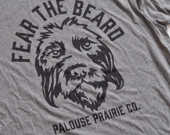 Może przedstawiać: Szara koszulka z czarnym nadrukiem przedstawiającym pysk psa w kółku z tekstem "Fear the Beard" i "Palouse Prairie Co."
