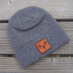 May include: Gray knit beanie with a brown leather patch featuring a deer head silhouette.