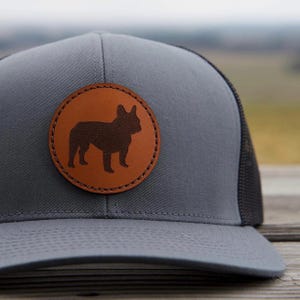 May include: A gray and black trucker hat with a brown leather patch featuring a silhouette of a French Bulldog.