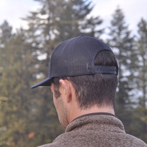 K&ouml;nnte beinhalten: Eine schwarze Mesh-Trucker-Kappe mit schwarzem Schirm. Die Kappe wird r&uuml;ckw&auml;rts getragen.
