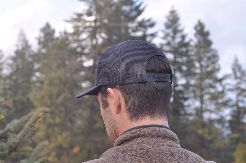K&ouml;nnte beinhalten: Eine schwarze Mesh-Trucker-Kappe mit schwarzem Schirm. Die Kappe wird r&uuml;ckw&auml;rts getragen.