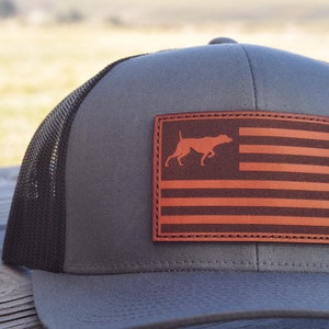 May include: A gray and black trucker hat with a brown leather patch featuring a silhouette of a dog and an American flag design.