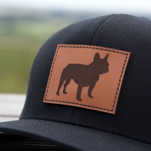 Puede incluir: Gorra de b&eacute;isbol negra con un parche de cuero marr&oacute;n que muestra la silueta de un bulldog franc&eacute;s. El parche rectangular est&aacute; cosido a la gorra. La gorra tiene una visera curva y una corona estructurada.